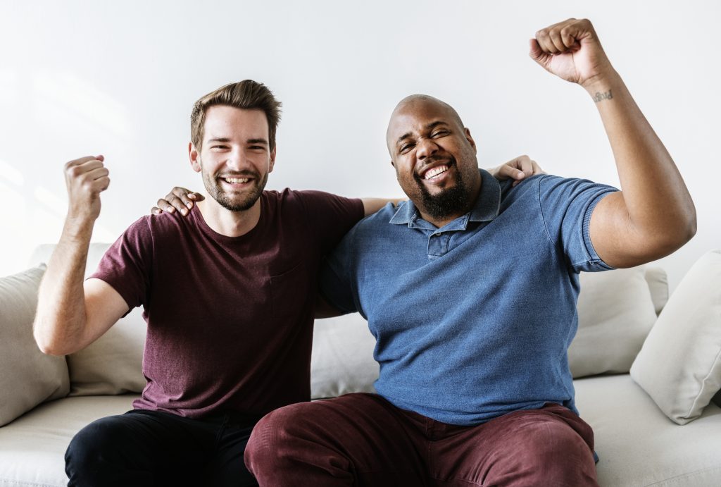 Two men sitting on a couch with their arms raised, appearing relaxed and engaged in conversation.