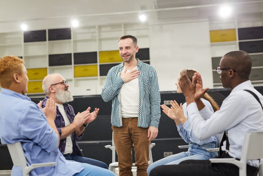 Portrait of smiling mature man introducing himself during therapy session in support group to people clapping, copy space