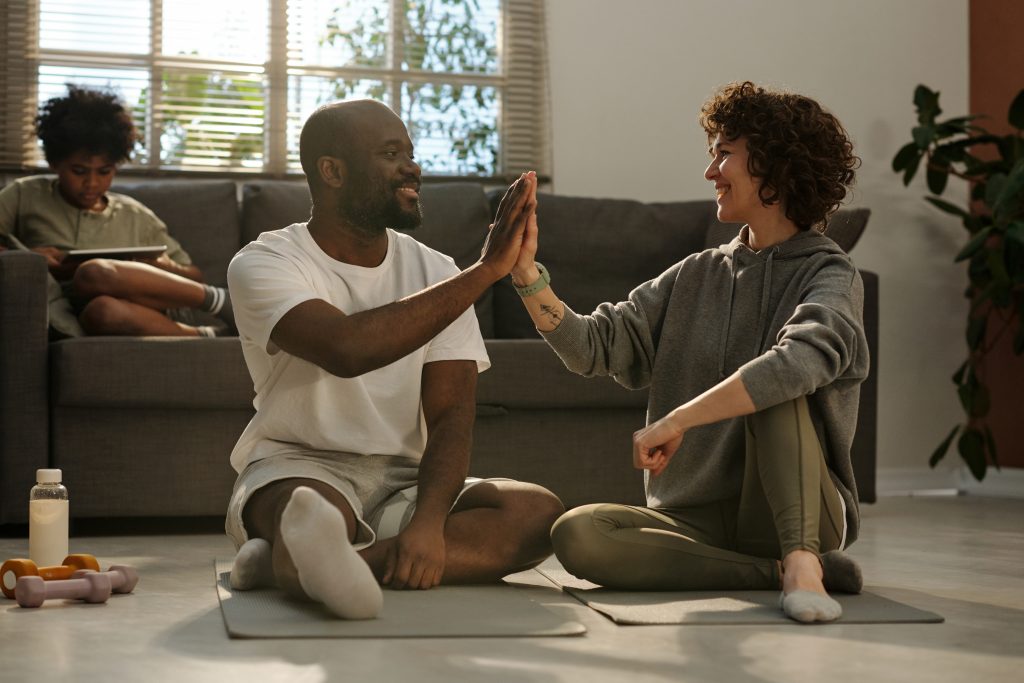 A man and woman sit on the floor, smiling with their hands raised in a joyful gesture.