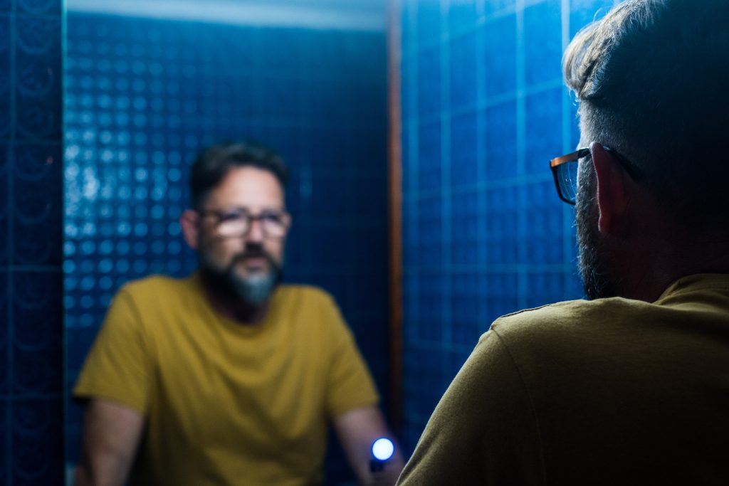 A man with glasses gazes into a mirror, reflecting on his appearance.