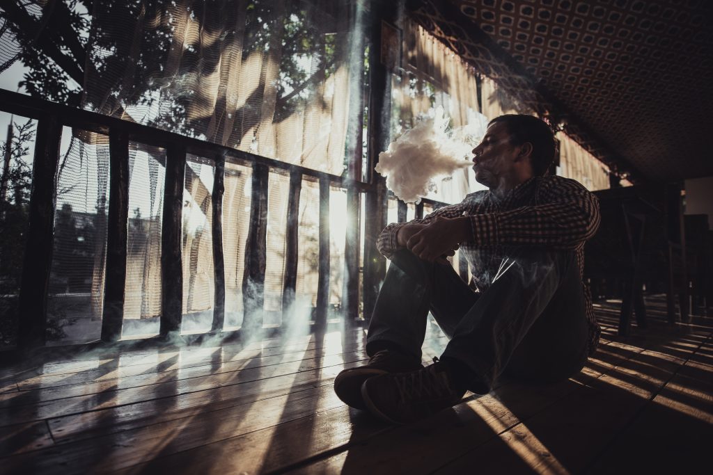 A man sits on a wooden floor, exhaling smoke, creating a mysterious atmosphere around him.